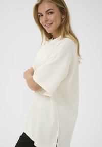 Mosolygó nő hosszú szőke hajjal, túlnőtt, off-white rövid ujjú felsőben és fekete nadrágban, karba tett kézzel áll a fehér háttér előtt.