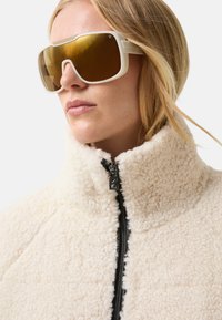 Kobieta nosząca duże białe okulary przeciwsłoneczne w oprawkach oraz zapinaną na zamek kremową kurtkę z polarowego materiału z wysokim kołnierzem.