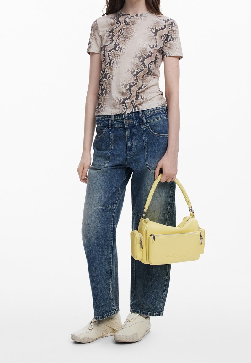 Femme portant un haut manches courtes imprimé serpent, jean bleu ample, baskets beiges, tenant un sac à main jaune avec poches zippées et anse tressée.