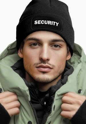 Black knit beanie with "SECURITY" label, worn over a hooded green jacket; layered textures and monochrome color scheme.