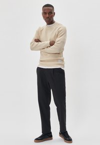 Pulover bej cu dungi, cu gât rotund, purtat peste un tricou alb. Pantaloni negri tapered și adidași negri cu talpă din cauciuc.