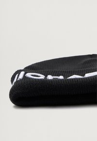 Zwarte gebreide beanie met een opgerolde zoom, voorzien van witte geborduurde tekst aan de voorkant. De textuur is geribbeld en zacht, geschikt voor koud weer.