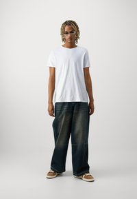 T-shirt branca de algodão, calças de ganga escuras de corte largo com um acabamento desbotado e ténis castanhos com atacadores brancos. Design simples.