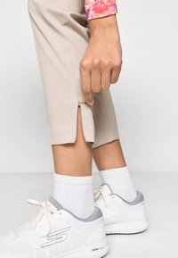 Κρεμ κολακευτικά παντελόνια με φερμουάρ στο τελείωμα, συνδυασμένα με λευκά αθλητικά παπούτσια και ριγωτές λευκές κάλτσες. Λείο ύφασμα, μινιμαλιστικός σχεδιασμός.