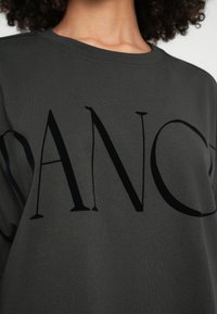 Tmavě šedá mikina s velkým černým nápisem "DANCE." Látka vypadá měkká s hladkou texturou.