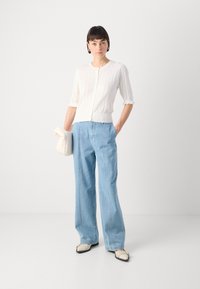 Cardigan bianco a maniche corte con motivo testurizzato, abbinato a jeans a gamba larga azzurri e ballerine bicolori. Tiene una borsa crema.