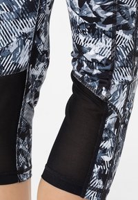 Zbliżenie na nogi w czarne i szare wzorzyste legginsy sportowe z czarnymi siateczkowymi panelami na łydkach.