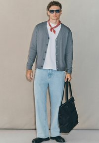 Tânăr purtând ochelari de soare negri, un cardigan gri, o cămașă albă, un fular roșu, blugi albaștri deschiși și pantofi negri, ținând o geantă de umăr neagră.