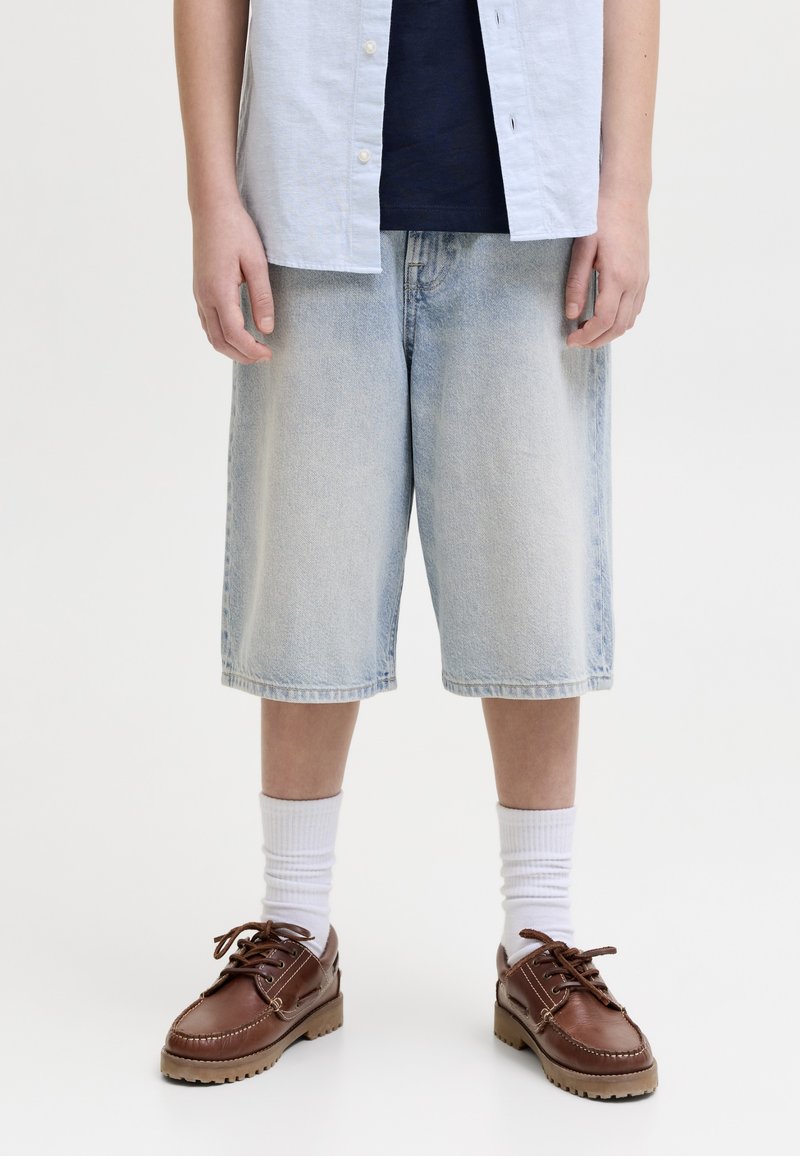 Personne portant un short en jean bleu clair jusqu'aux genoux, des chaussettes blanches, des chaussures en cuir marron à lacets, une chemise bleu marine et une chemise bleu clair boutonnée.