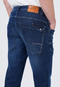 Donkerblauwe denim jeans met een gestructureerde afwerking, voorzien van een leren taillestrik, vijf zakken en contrasterende stiksels.