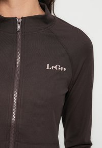 Σκούρο καφέ μπουφάν με φερμουάρ, με ραβδωτή υφή, ψηλό γιακά και ροζ λογότυπο "LeGer" στην αριστερή πλευρά. Διαθέτει ασημένιο φερμουάρ.