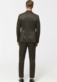 Man standing with back facing camera, wearing a dark brown suit and black dress shoes against a plain light background.