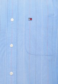 Jasnoniebieska koszula z guzikami, wykonana z tkaniny tkanej, z pionowymi białymi i czerwonymi paskami oraz małym haftowanym logiem na kieszeni piersiowej.