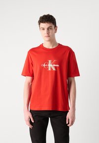 Crvena pamučna majica s kratkim rukavima i okruglim izrezom, s velikim bijelim i srebrnim printanim logotipom na prednjoj strani.