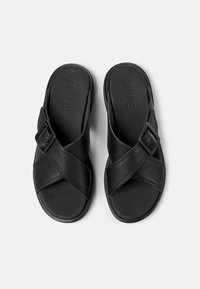 Black leather sandals with a cross-strap design, featuring a buckle accent and a flat sole. Textured insole with embossed brand name.