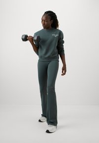 Zelená mikina s bielym logom, priliehavé zelené nohavice a šedé atletické topánky. Drží čiernu činku. Jednoduchý, atletický dizajn.