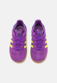 Lila mockaim sportskor med gula ränder, platta snören och en blå hälaccent; med en texturerad gummisula.
