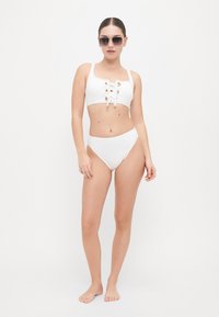 Witte bikiniset met een cropped top met veters en gouden oogjes, gecombineerd met hooggelegen onderstukken. Model draagt een donkere zonnebril.