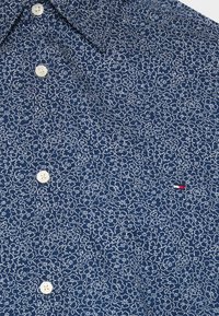 Camicetta blu navy con un motivo floreale bianco. Presenta una tasca piccola con un accento rosso, bianco e blu. Realizzata in tessuto testurizzato.
