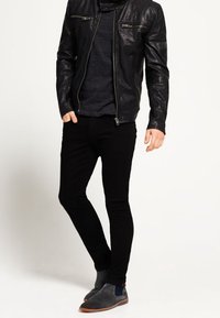 Giacca in pelle nera con accessori in argento, maglione scuro, pantaloni neri slim-fit e stivali in suede grigi con accenti blu sulla tomaia.