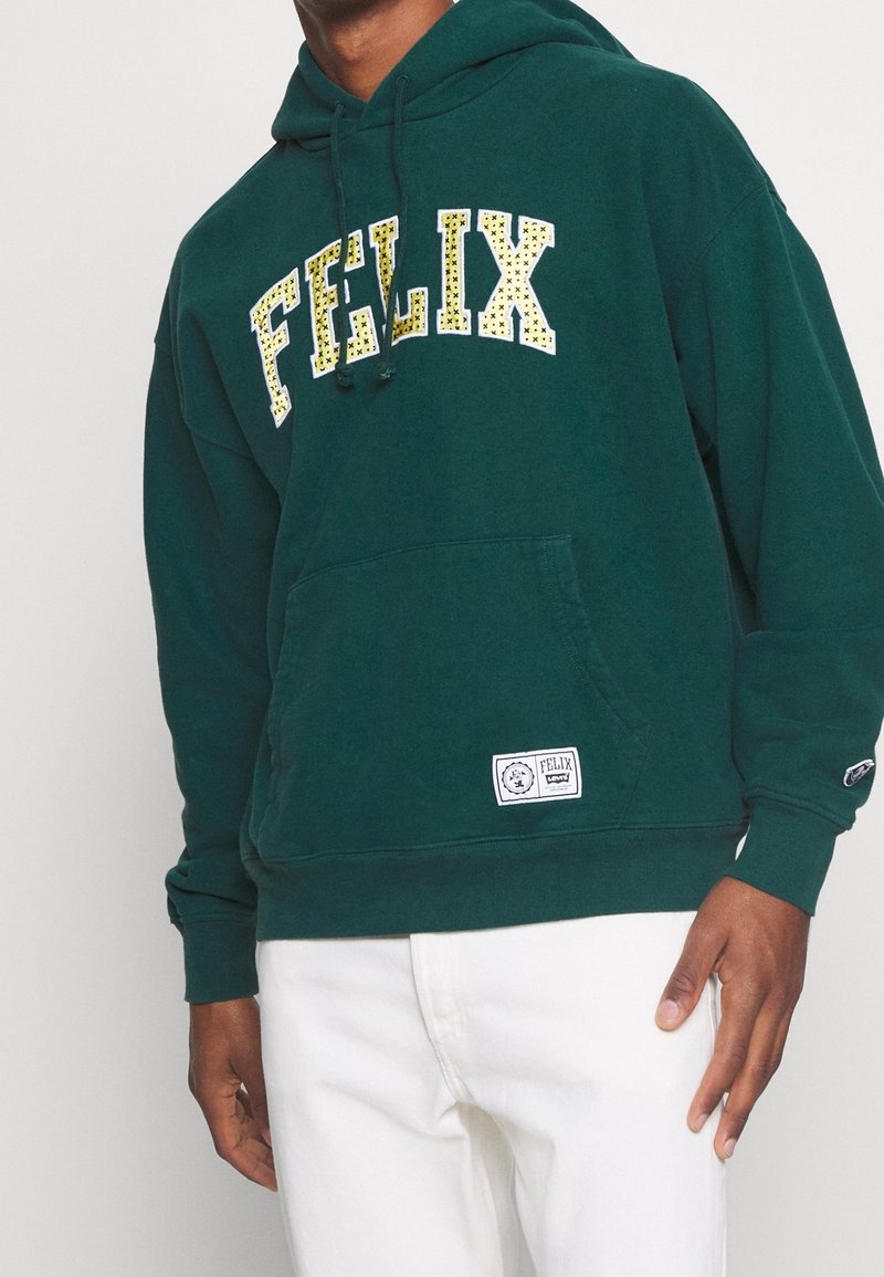 Hombre con sudadera con capucha verde oscuro con el texto "FELIX" en un patrón de estrellas amarillas y pantalones blancos, de pie contra un fondo claro y liso.