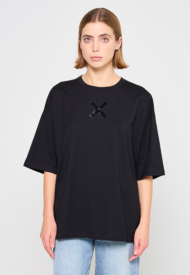 Femme aux cheveux blonds courts portant un t-shirt noir surdimensionné avec un X noir brillant sur la poitrine et un jean bleu clair, se tenant devant un fond blanc.