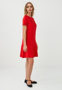 Une femme se tient de profil, portant une robe rouge vif à volants et des ballerines noires, sur un fond blanc uni.