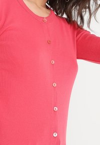 Pink ribbed cardigan with round neckline, featuring a row of light buttons along the front and long sleeves. Smooth texture, fitted design.