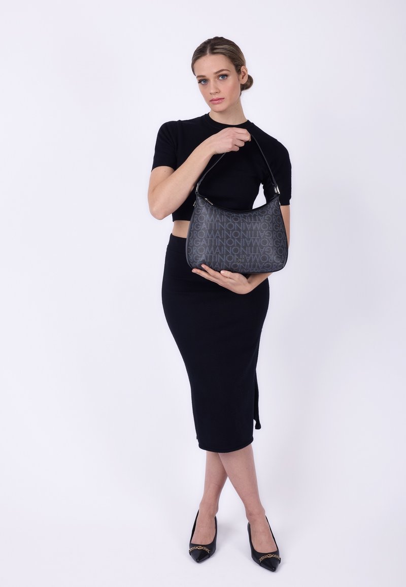 Donna in abito nero che tiene una borsa firmata nera con un motivo testuale davanti a uno sfondo bianco.