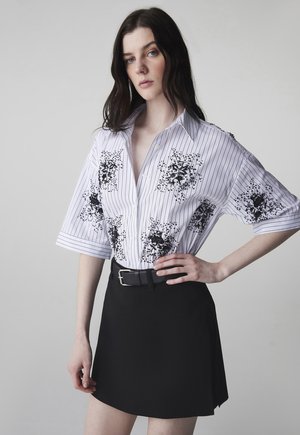 Vrouw met lang donker haar, gekleed in een wit kort mouwen overhemd met zwarte bloemenpatronen en een zwarte rok met een riem.