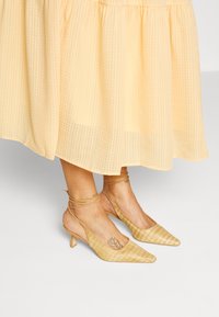 Sapatos de salto agudo amarelo-claro com um acabamento têxtil texturizado, apresentando uma tira fina no tornozelo e um pequeno salto bloco.