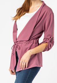 Veste légère bordeaux enveloppante avec une coupe décontractée, taille nouée et manches retroussées agrémentées de boutons. Texture lisse, design décontracté.