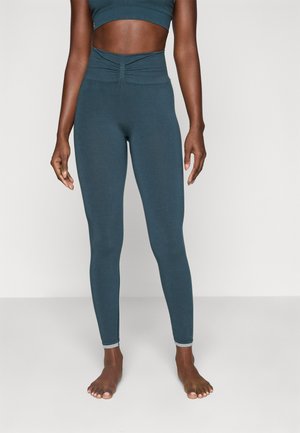 Woman wearing high-waisted, ruched dark teal leggings and matching cropped athletic top, standing barefoot against a plain light background.