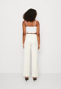 Vrouw met krullend haar die een witte crop top, crèmewitte hoge taille broek en zwarte hakken draagt, draaiend naar een simpele muur.