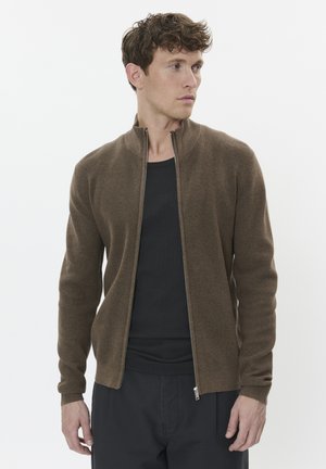 Jeune homme portant un pull marron zippé par-dessus une chemise noire, regardant vers sa gauche sur un fond blanc uni.