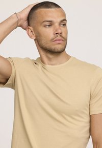 T-shirt beige à manches courtes en matière douce ; présente un col rond, des coutures minimales, une texture lisse et une coupe décontractée.
