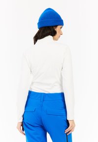 Witte longsleeve met hoge kraag, gecombineerd met felblauwe broek met zakken en een gladde textuur. De outfit wordt compleet gemaakt met een blauwe gebreide beanie.
