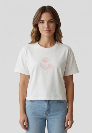 Giovane donna che indossa una t-shirt corta bianca con illustrazioni di conchiglie rosse e jeans azzurri, in piedi davanti a uno sfondo grigio uniforme.