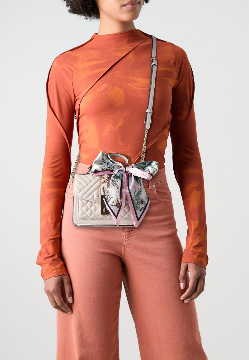 Sac à bandoulière matelassé de couleur gris clair avec une écharpe rose à motif floral décoratif attachée à la poignée. Il possède une chaîne comme sangle et des éléments en métal.