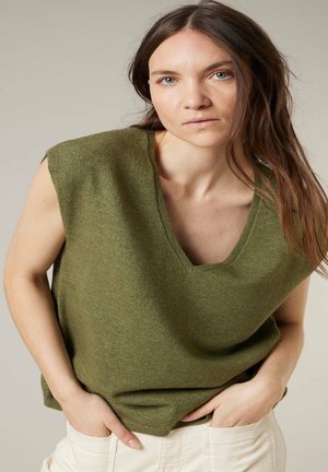 Jeune femme aux longs cheveux bruns portant un haut en tricot sans manches vert olive et un pantalon blanc, posant les mains dans les poches devant un fond uni.