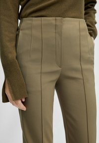 Pantalon kaki à la coupe ajustée, doté de plis à l'avant, de poches latérales et d'un tissu lisse et texturé. La ceinture est plate, sans fermetures visibles.