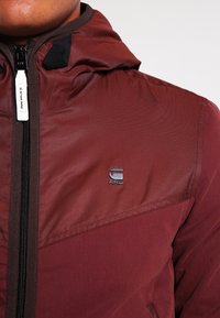Röd tygjacka med huva, med dragkedjestängning, texturerade övre paneler och en svart logotyp på bröstområdet. Slätt och hållbart material.