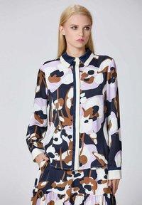 Chemisier à manches longues et col, avec un motif floral bleu marine, blanc et marron sur un fond lilas clair. Tissu lisse avec fermeture à boutons.