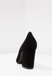 Scarpa donna nera in suede con tacco spesso, fotografata su uno sfondo bianco dalla parte posteriore sinistra.