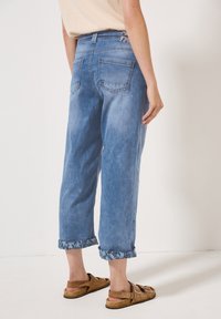 Person som bär ljusblå, uppvikta, korta jeans och bruna mockasandaler står mot en enkel vit bakgrund.