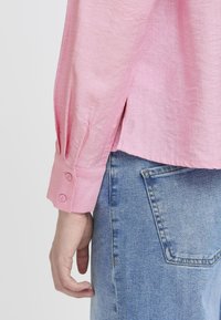 Rosa Langarmhemd aus strukturiertem Stoff mit Knopf-Detail an den Manschetten, kombiniert mit hellblauen Jeans, die eine Gesäßtasche zeigen.