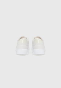 Une paire de baskets blanches basses vue de l'arrière, présentant des tiges lisses et des semelles en caoutchouc blanches épaisses sur un fond clair.