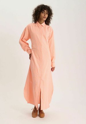 Femme aux cheveux bouclés portant une longue robe chemise à carreaux pêche et des chaussures marron à lacets, debout devant un fond uni.