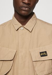 Beige button-up shirt made of textured fabric, featuring two chest pockets and a black label with yellow text on the right pocket.