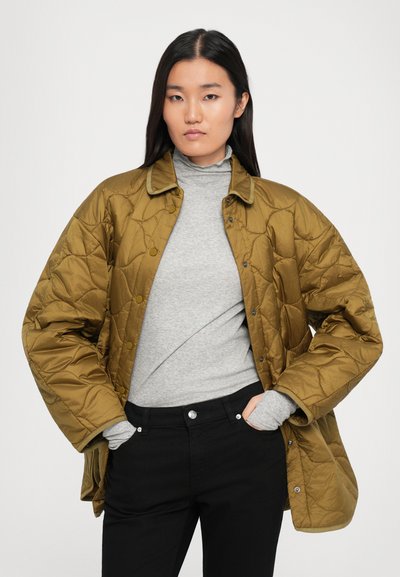 Sakrākta olīvzaļa jaka ar apkakles izgriezumu, kas aprīkota ar slēdzamām pogām un brīvu piegriezumu, uzvilkta uz pelēka džemperi ar augstu apkakli un melniem biksēm.